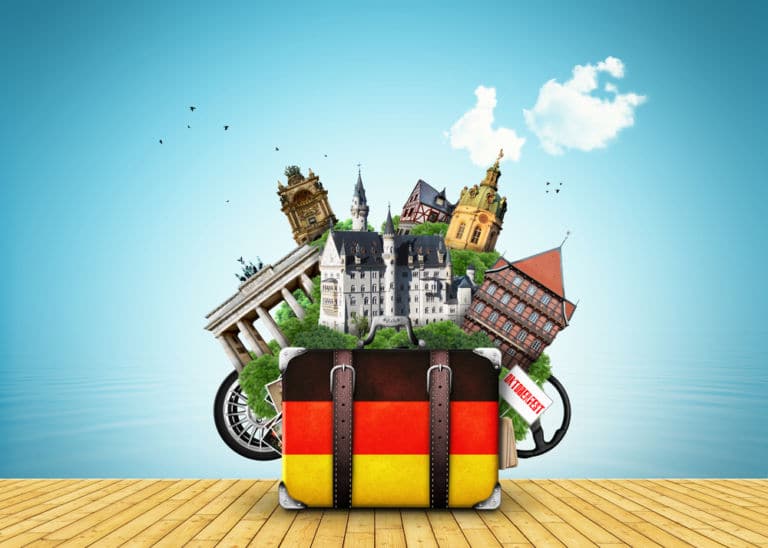 Im Bild ist eine Truhe von vorne zu sehen die wie angemalt wurde wie eine Deutschlandfahne. Im Hintergrund sind Wahrzeichen Deutschlands zu sehen, darunter das Brandenburger Tor und Schloss Neuschwanstein, die sich unter anderem in der gepackten Truhe befinden können als Titelbild zur Folge 7 – Wenn sich zwei Deutsche das Wasser reichen
