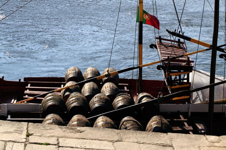 Das Bild zeigt ein traditionelles portugiesisches Boot, eine sogenannte Rabelo-Barkasse, beladen mit Fässern auf dem Fluss Douro in Porto, Portugal. Das Titelbild zur Folge 44 – Mit allen Fässern gewaschen