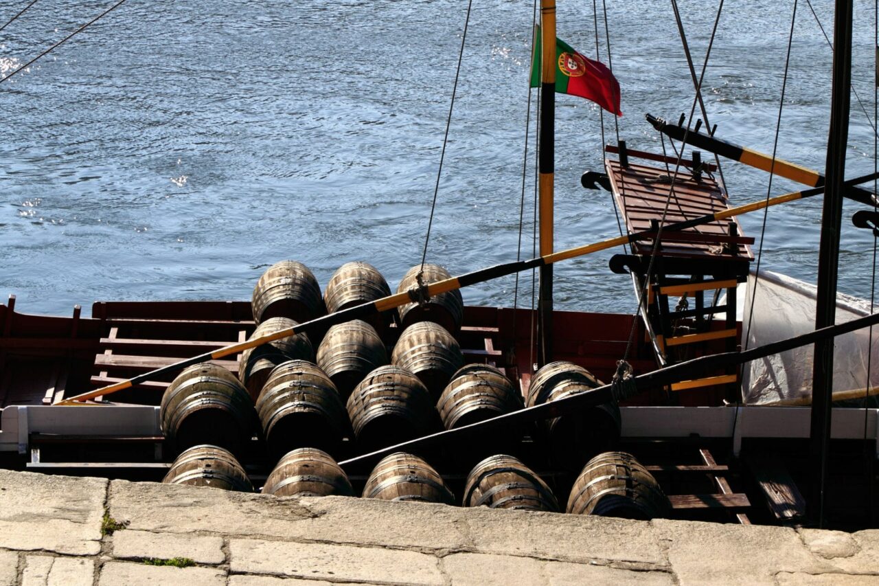Das Bild zeigt ein traditionelles portugiesisches Boot, eine sogenannte Rabelo-Barkasse, beladen mit Fässern auf dem Fluss Douro in Porto, Portugal. Das Titelbild zur Folge 44 – Mit allen Fässern gewaschen