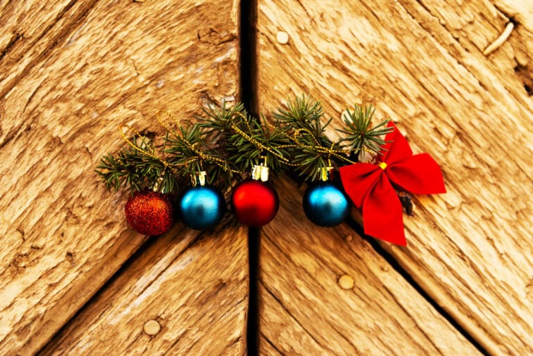 Zwei blaue und zwei rote Weihnachtskugeln mit goldenem Band und einer roten Schleife die an einem Tannenzweig auf hölzernem Grund liegen als Titelbild zur Folge 64 – Heiße Weihnacht: Rückblick mit Punsch