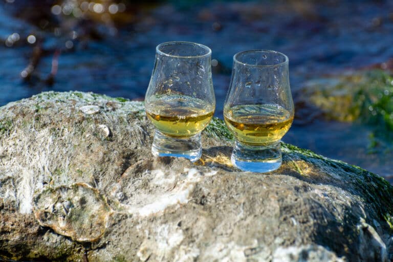 Zwei mit Whisky gefüllte Glencairn Gläser auf einem Stein an einem Fluss als Titelbild zur Folge 71 – Süße Versuchungen von GlenAllachie