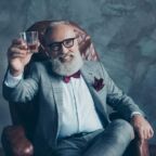 Portrait of bearded, lucky, old rich man in formal wear with bow tie and pocket square, sitting on chair, holding, raise glass with whiskey, crypto-currency, shares, stock