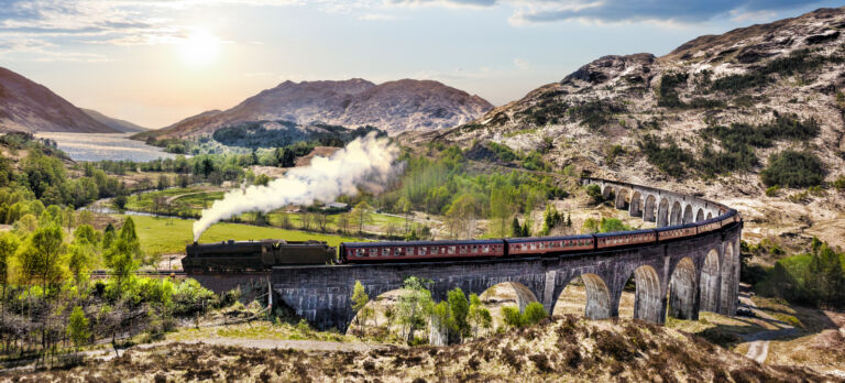Glenfinnan-Eisenbahnviadukt in Schottland mit dem Jacobite-Dampfzug vor dem Sonnenuntergang über dem See. Das Titelbild zur Folge 93 – Reise durchs gelobte Land
