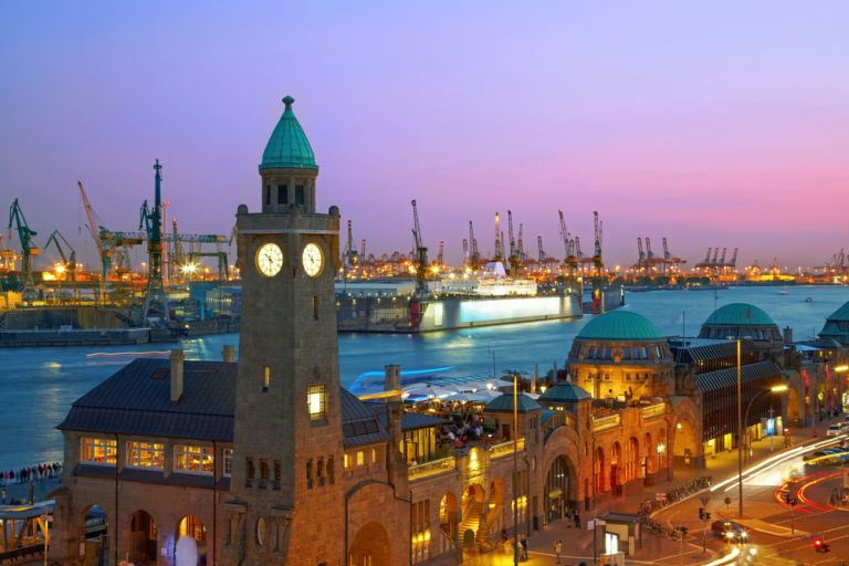 Abendstimmung an den Landungsbrücken in Hamburg mit Blick auf Hafen, Kräne und Schiffe – leuchtende Stadtlichter, Uhrturm im Vordergrund, realistischer Fotostil. – Titelbild Folge 130 – Der versierte Braumeister