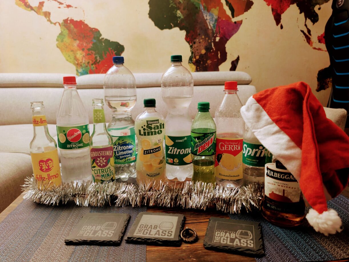 Titelbild zur Folge 202 'Weihnachtszitronenlimoinferno'. Auf einem Tisch steht eine Flasche Kilbeggan Irish Whiskey inmitten eines chaotischen Meeres aus verschiedenen Zitronenlimonaden-Flaschen (Sprite, Fritz, Mountain Dew, Gerolsteiner etc.)