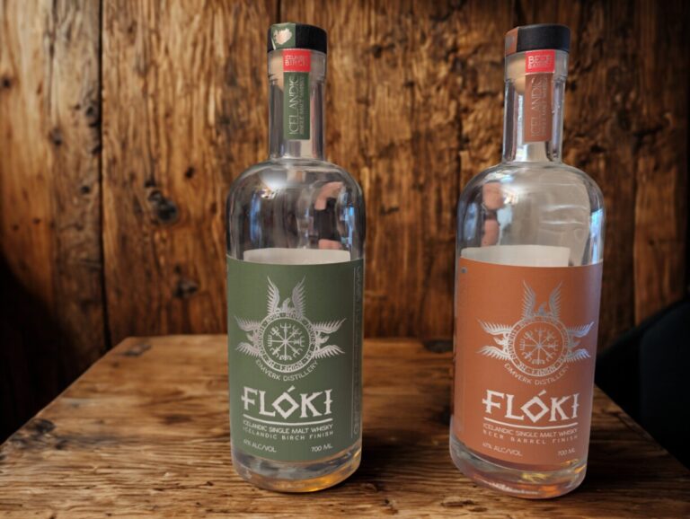 Zwei Flaschen Flóki Icelandic Single Malt Whisky stehen nebeneinander auf einem massiven Holztisch vor einer rustikalen Holzwand. Links steht die Flasche mit dem olivgrünen Etikett (Icelandic Birch Finish), rechts daneben die Flasche mit dem kupferfarbenen Etikett (Beer Barrel Finish).