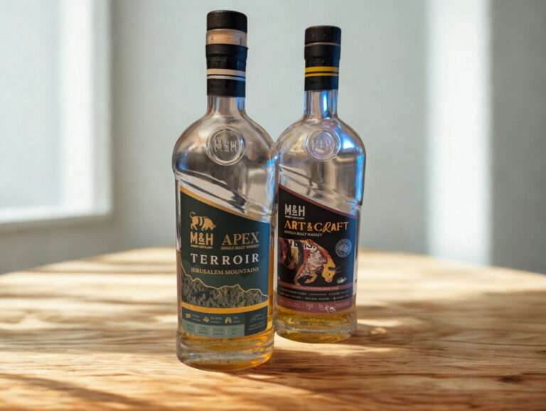 Zwei Whiskyflaschen der israelischen Destillerie Milk & Honey (M&H) stehen nebeneinander auf einem Tisch vor einer schlichten weißen Wand. Links steht der 'M&H Apex Terroir Jerusalem Mountains' mit einem hellen Etikett, rechts daneben der 'M&H Art & Craft' mit dem bunten, gezeichneten Stier-Motiv auf dem Label.