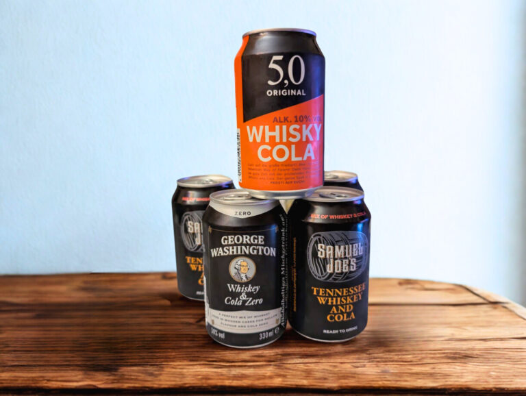 Ein Turm aus RTD-Whisky-Cola-Dosen ('Alufässchen voll Abwasser') steht auf einem Holztisch vor einer weißen Wand aus Folge 210. Unten stehen zwei Dosen: 'Samuel Joe's Tennessee Whiskey & Cola' und 'George Washington Whiskey & Cola Zero'. Oben in der Mitte balanciert die einzelne Dose '5,0 Original Whisky Cola'. Dahinter wiederholen sich die 'Samuel Joe's' und 'George Washington' Dosen nebeneinander, aber ohne die 5,0er on top.