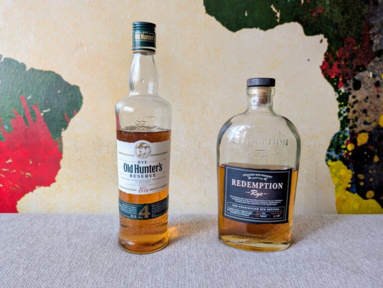 Zwei Whiskyflaschen stehen nebeneinander auf einer hellgrauen Stoffoberfläche vor einer Wand mit einer bunten Weltkarten-Bemalung. Links steht die hohe, schmale Flasche des tschechischen 'Old Hunter's Reserve Rye' mit grünem Schraubverschluss. Rechts daneben steht die markante, breitere Flasche des amerikanischen 'Redemption Rye' mit schwarzem Korken.