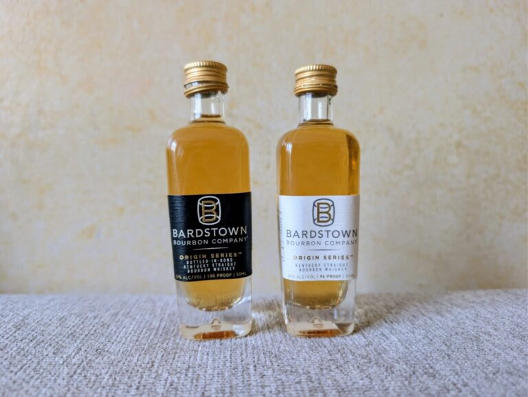 Zwei 50ml-Miniaturflaschen der Bardstown Bourbon Company stehen nebeneinander auf einer hellen Stoffoberfläche. Links die "Bottled-in-Bond"-Edition mit schwarzem Etikett, rechts die "High Rye"-Edition mit weißem Etikett. Beide Flaschen haben goldene Schraubverschlüsse und zeigen das bernsteinfarbene Destillat.