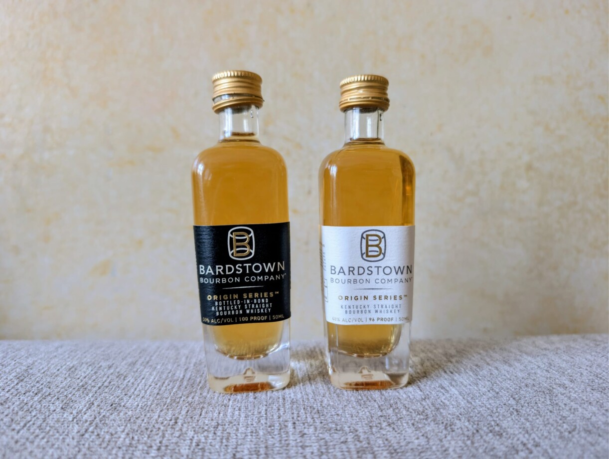 Zwei 50ml-Miniaturflaschen der Bardstown Bourbon Company stehen nebeneinander auf einer hellen Stoffoberfläche. Links die "Bottled-in-Bond"-Edition mit schwarzem Etikett, rechts die "High Rye"-Edition mit weißem Etikett. Beide Flaschen haben goldene Schraubverschlüsse und zeigen das bernsteinfarbene Destillat.