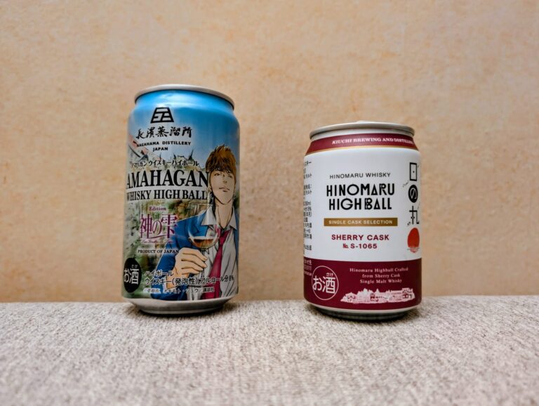 Zwei japanische Whisky-Highball-Dosen stehen nebeneinander auf einem hellen Stoffuntergrund vor einer strukturierten beigen Wand. Links steht die etwas größere, hellblaue Amahagan-Dose der Nagahama Distillery, die einen jungen Mann mit Weinglas im Manga-Stil ("Drops of God") zeigt. Rechts daneben steht die etwas kleinere, weiß-rote Hinomaru-Dose von Kiuchi Brewing and Distilling (Sherry Cask Edition No. S-1065) mit einem kleinen roten Sonnen-Symbol auf der rechten Seite.