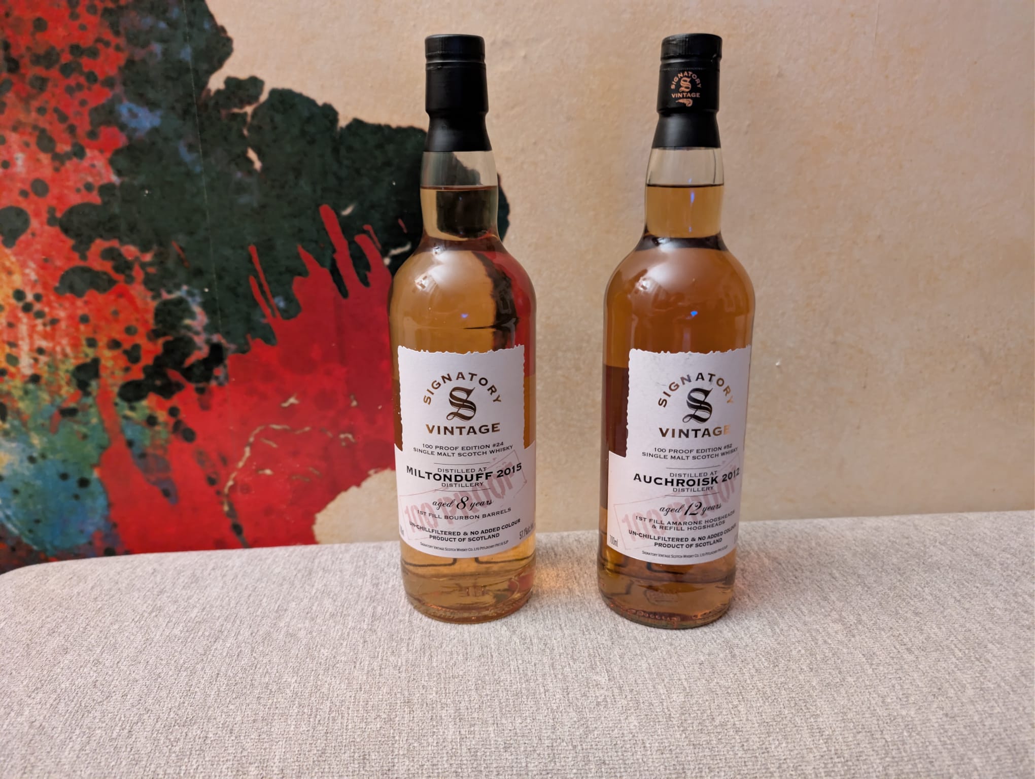 Zwei Whiskyflaschen des unabhängigen Abfüllers Signatory Vintage aus der 100 Proof Edition stehen nebeneinander. Links die Flasche Miltonduff 2015 (8 Jahre, First Fill Bourbon Barrels) und rechts die Flasche Auchroisk 2012 (12 Jahre, 1st Fill Amarone & Refill Hogsheads). Im Hintergrund ist ein farbenfrohes Kunstwerk mit roten und blauen Farbelementen zu sehen.